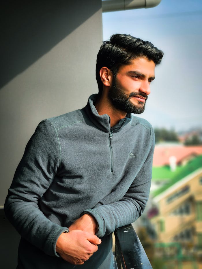 about-us A relaxed man in a gray sweater enjoys a sunny day outdoors, leaning on a railing.